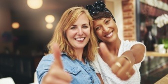 Two young women giving thumbs up with brick building in background